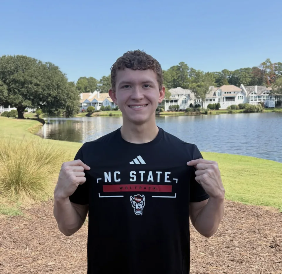 Charlie Camplin holding up an NC State Wolfpack shirt beside a lake